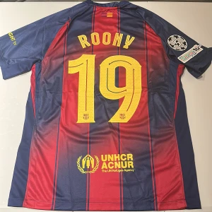 FC Barcelona matchtröja Nike Roony 19 - Säljer en officiell FC Barcelona matchtröja från Nike med Roony och nummer 19 på ryggen. Tröjan är blå och röd med gula detaljer, randigt mönster, Champions League-märke på ärmen och UNHCR samt Spotify-logga. Tillverkad i lätt och ventilerande polyester. Replika, endast handtvätt gäller!