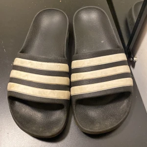 Svarta Adidas sandaler med vita ränder - Klassiska svarta Adidas sandaler med tre vita ränder över foten. Tillverkade i syntetmaterial med platt sula och öppen tå. Perfekta för sommaren eller att slappa i hemma. Adidas-logga på sulan och sidan.