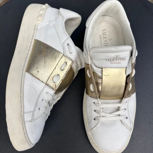 Valentino Rockstud vita sneakers - Snygga vita Valentino Rockstud sneakers med breda guldiga band och ikoniska nitar. Skorna har snörning, platt sula och är tillverkade i skinn. Perfekta för dig som vill sticka ut med en lyxig och edgy vibe.