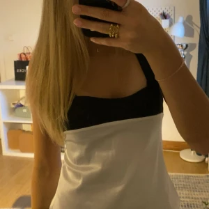 Svartvit satinlinnetopp med breda axelband - Säljer en snygg svartvit linne-topp i satin med breda svarta axelband och fyrkantig urringning. Toppen har en stilren och modern look med kontrast mellan svart upptill och vitt nedtill. Perfekt för dig som gillar minimalistisk stil och vill sticka ut.