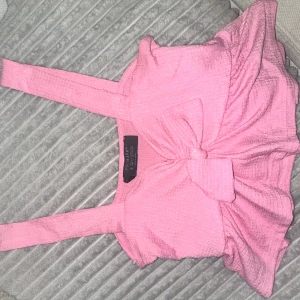 Rosa linnetopp med rosett  - Supersöt rosa linnetopp från med breda axelband och en stor rosett framtill. Toppen har en lätt vågig nederkant och är gjord i ett mjukt, strukturerat tyg som känns skönt mot huden. Perfekt för att lyfta vilken outfit som helst!