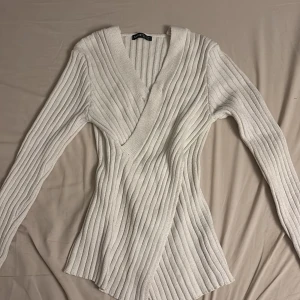 Vit omlott tröja från SHEIN - Säljer en vit ribbad omlott tröja från SHEIN. Tröjan har lång ärm, markerad midja och djup v-ringning. Den är stickad i mjukt material och passar perfekt för lager-på-lager-stil. Enkel att matcha med jeans eller kjol.