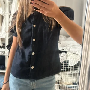 Mörkblå kortärmad jeans tröja med knappar - Säljer en mörkblå jeans skjorta i bomull med korta puffärmar och silvriga knappar framtill. Skjortan har en rak passform och klassisk krage, perfekt för en chill och stilren look. Enkel att matcha med jeans eller kjol.