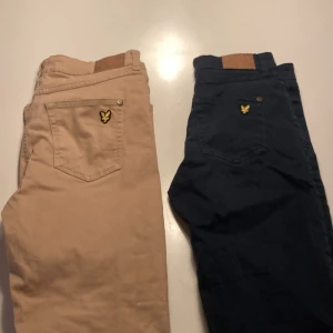 Lyle & Scott chinos i beige och marinblå - Två par snygga chinos från Lyle & Scott Junior, ett par i beige och ett par i marinblått. Klassisk femficksmodell med gylf och knapp, samt det ikoniska Lyle & Scott-märket på bakfickan. Tillverkade i mjuk bomull för en skön känsla.