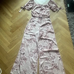 Blommig byxdress från Gina Tricot - Supersöt ljusrosa byxdress från Gina Tricot med blommigt mönster i bruna toner. Vida ben och off-shoulder topp med puffärmar och knytband. Mjuk, glansig känsla i materialet som ger en lyxig vibe. Perfekt för dig som vill sticka ut med en feminin look.