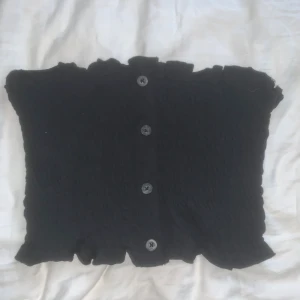 Svart bandeau topp  - Svart bandeau topp från Brave Soul med smockad struktur och dekorativa knappar framtill. Toppen har volangkant upptill och nedtill för en söt touch. Perfekt att styla till jeans eller kjol för en trendig look.