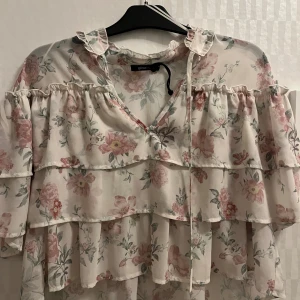 Blommig volangblus från Gina Tricot - Supersöt blommig blus i ljusbeige med rosa och gröna detaljer. Blusen har flera volanger, v-ringning med knytband och korta ärmar. 