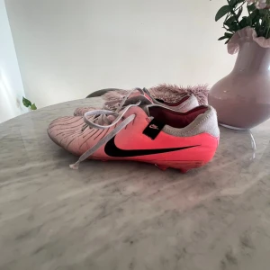 Nike rosa fotbollsskor med snörning - Säljer ett par snygga Nike fotbollsskor i rosa och grått med svart swoosh på sidan. Skorna har snörning och är designade för att ge bra grepp på planen. Ovandelen är i syntetmaterial och hälen är klädd i grått tyg. Perfekta för dig som vill sticka ut på matchen!