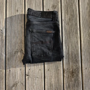 Svarta jeans från Nudie Jeans, 31/34 - Snygga svarta jeans från Nudie Jeans i klassisk rak modell. Jeansen har fem fickor, svarta sömmar och diskret logga på bakfickan. Tillverkade i mjukt denimtyg som sitter skönt och passar till många olika stilar. Grim Tim 31/34