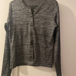 Gråmelerad kofta från Sisters Point - Gråmelerad kofta från Sisters Point i storlek M/L. Koftan har rund halsringning, långa ärmar och knäppning framtill med svarta knappar. Materialet är mjukt och stretchigt, perfekt för lager-på-lager. Snyggt diskret mönster som passar till allt.