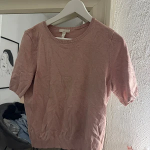 Ljusrosa stickad topp från H&M - Säljer en ljusrosa stickad topp från H&M med rund halsringning och korta ärmar. Toppen har ribbade muddar vid hals, ärmslut och nederkant. Enkel och stilren modell som är lätt att matcha till olika outfits.