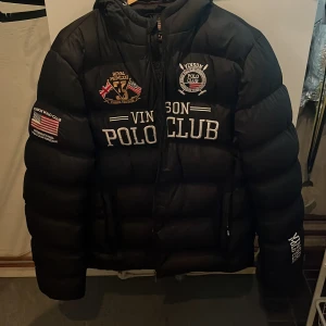 Svart pufferjacka Vinson Polo Club M - Svart pufferjacka från Vinson Polo Club med broderade loggor och patchar på bröstet och ärmen. Jackan har huva, dragkedja framtill och tydlig quiltad design. Perfekt för kalla dagar och riktigt snygg streetstil. Ny pris 1499