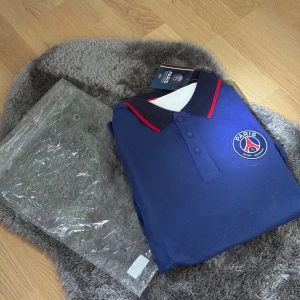 PSG pikétröja marinblå  - Marinblå pikétröja från Paris Saint-Germain med klubbens logga på bröstet. Tröjan har krage med röda detaljer och knäppning framtill. Perfekt för dig som älskar fotboll och vill visa ditt stöd för PSG.
