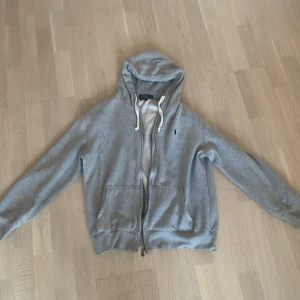 Grå hoodie från Polo Ralph Lauren - Säljer en grå hoodie från Polo Ralph Lauren med dragkedja och huva med vita snören. Klassisk logga broderad på bröstet och två fickor framtill. Perfekt för chill dagar eller när du vill ha en enkel och stilren look. DET STÅR XXL MEN KÄNNS SOM M (Har storlek M) skriv vid frågor!🤔🙋‍♂️🤝🏽                                                            Den är i gott skick förutom att Tjedjan gått (Ur led) men det kan man fixa inom 10 min hemma👌🏽