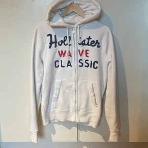 Vit hoodie från Hollister med tryck - Vit hoodie från Hollister med broderat tryck i marinblått och rött på framsidan. Tröjan har huva med snörning, två fickor fram och dragkedja. Mjuk insida i fleece och långärmad modell, perfekt för chill dagar.