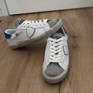 Philippe Model vita sneakers strl 43 - Snygga vita Philippe Model sneakers med grå mockadetaljer och blå hälkappa. Skorna har klassisk snörning, rund tå och är tillverkade i skinn och mocka. Sköldlogga på sidan och Philippe Model Paris-tryck på hälen. Perfekta för en clean och stilren look.