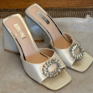 Vita satin klackar - Snygga klackskor i vit satin med fyrkantig tå och bred klack, storlek 38, från Asos. Skorna har en glittrande strassdetalj framtill som ger extra bling. Perfekta för dig som vill sticka ut med en elegant och trendig look.  För hög klack för mig och kommer drf inte till användning längre. Fortfarande i gott skick.