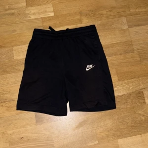 Svarta Nike shorts - Svarta shorts från Nike med vit logga på vänster ben. Modellen har elastisk midja med snörning för skön passform och är tillverkad i mjuk bomull. Perfekta för träning eller chill. Klassisk och enkel design utan mönster. Storleken är 147-158 cm eller L i barnstorlek