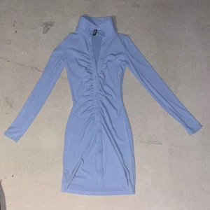 Ljusblå skjortklänning från H&M Divided - Supersnygg ljusblå kort skjortklänning från H&M Divided med lång ärm, markerad krage och knappar hela vägen framtill. Klänningen har rynkade detaljer längs knappraden och är gjord i ett mjukt, stretchigt material som sitter slimmat på kroppen.