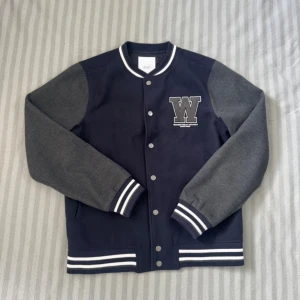 Mörkblå bomberjacka från Grunt S - Snygg mörkblå bomberjacka från Grunt med grå ärmar och vita ränder på muddar och krage. Jackan har tryckknappar framtill och en stor W-logga på bröstet. Tillverkad i 100% polyester, perfekt för dig som gillar college-stil.