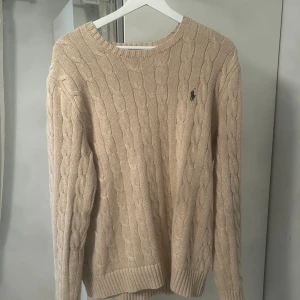 Beige kabelstickad tröja Polo Ralph Lauren - Kabelstickad beige tröja från Polo Ralph Lauren med rund halsringning och broderad logga på bröstet. Tröjan har ribbade muddar vid ärmslut och nederkant. Perfekt under höst och vinter. Passar även L väldigt bra!