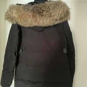 Svart dunjacka med fuskpäls från Cecion - Svart dunjacka från Cecion Sweden i storlek 36. Jackan har en stor, fluffig fuskpäls runt huvan, flera dragkedjeförsedda fickor och snygga detaljer i svart. Perfekt för kalla dagar med sin långa modell och varma känsla.