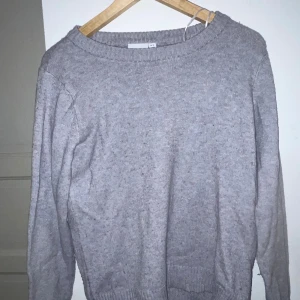 Grå stickad tröja från Vila - Mysig grå stickad tröja från Vila med rund halsringning och ribbade muddar vid ärmslut och nederkant. Perfekt för lager-på-lager och enkel att matcha med både jeans och kjol. Klassisk modell med normal passform och långa ärmar.