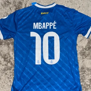 Real Madrid Mbappé 10 blå Adidas tröja - Snygg blå Real Madrid fotbollströja från Adidas med Mbappé och nummer 10 på ryggen. Tröjan har vita detaljer, klubbmärke på bröstet, sponsortryck och klassiska Adidas-ränder på axlarna. Tillverkad i lätt och ventilerande material, perfekt för match eller träning.
