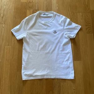 Vit Moncler t-shirt - Clean vit t-shirt från Moncler med klassisk passform och korta ärmar. Diskret Moncler-logga på bröstet ger en exklusiv vibe. Tillverkad i mjuk bomull som känns skön mot huden. Perfekt för dig som gillar stilrena och lyxiga basplagg.