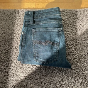 Mörkblå skinny jeans från Nudie Jeans - Säljer ett par mörkblå skinny jeans från Nudie Jeans i storlek 28/30. Klassisk femficksmodell med broderad bakficka och smal passform. Jeansen har normal midja och är tillverkade i stretchig denim för en bekväm känsla. Priset e ej hugget i stenen