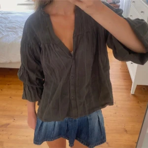 Grå/khaki blus med vida ärmar - Säljer en blus med v-ringning och knappar framtill. Blusen har vida, trekvartsärmar och är tillverkad i ett lätt, luftigt material som ger en avslappnad känsla. Perfekt för dig som gillar en chill och stilren look.