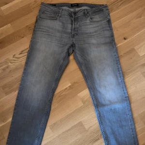 Grå tapered jeans storlek 34/32 - Snygga grå jeans med tvättad look och tapered passform. Klassisk femficksmodell med normal midja och raka ben som smalnar av. Jeansen är i mjukt denimtyg och passar perfekt till en avslappnad stil.
