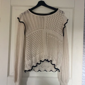 Beige stickad tröja med volang Vero Moda - Superfin beige stickad tröja från Vero Moda med lång ärm och coola svarta detaljer vid hals, axlar och nederkant. Tröjan har olika stickade mönster och volangkant som ger en unik look. Perfekt för dig som gillar detaljer och vill ha något extra i garderoben.