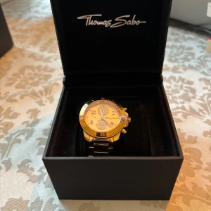 Thomas Sabo roséguldklocka dam - Snygg damklocka från Thomas Sabo med boett och armband i roséguldtonat stål. Urtavlan har glittriga detaljer, arabiska siffror och självlysande visare. Diameter 40 mm, tjocklek 13 mm. Mineralglas och viklås. Kvartsurverk med datumfunktion.