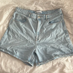 Ljusblå jeansshorts från Zara - Säljer ett par ljusblå jeansshorts från Zara med klassisk femficksdesign och hög midja. Shortsen har bälteshällor, knapp och dragkedja framtill samt rå kant nedtill för en chill vibe. Säljer då de är för stora för mig och inte längre kommer till användning.