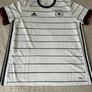 Tysklands vit randig fotbollströja XL - Snygg vit fotbollströja från Adidas med svarta horisontella ränder och Tysklands landslagsmärke på bröstet. Kortärmad modell med svart krage och detaljer i rött, svart och guld på ärmsluten. Tillverkad i lätt och ventilerande Aeroready-material.