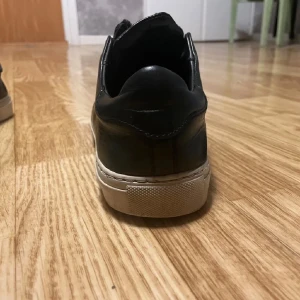 Svarta sneakers Axel Arigato med dragkedja - Säljer ett par svarta sneakers från Axel Arigato med stilren design och dragkedja framtill istället för klassisk snörning. Skorna är gjorda i skinn med vit platt sula och har en rund tå. Medföljer originalkartong och dustbag.