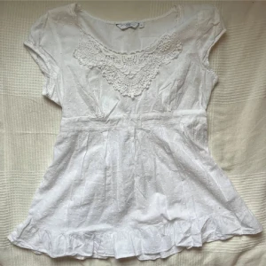 Vit blus med spets och volang - Supersöt vit blus med spetsdetaljer vid halsen, små prickar i tyget och volang längst ner. Knytband i midjan för en snygg passform och korta ärmar. Perfekt för en romantisk och fräsch look.