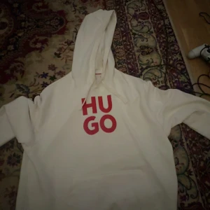Vit hoodie från Hugo med röd logga - Säljer en vit hoodie från Hugo med stor röd logga på bröstet. Tröjan har huva med snörning och en klassisk känguruficka framtill. Perfekt för dig som gillar stilrena och bekväma plagg med tydlig branding.