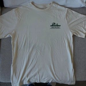 Vit t-shirt från Aimé Leon Dore - Vit t-shirt från Aimé Leon Dore med grönt tryck på bröstet och stort grafiskt tryck på ryggen. Klassisk passform med rund hals och korta ärmar. Tillverkad i mjuk bomull, perfekt för en clean och stilren look. Jättefint skick!! Nypris 800kr