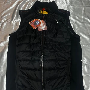 Svart dunväst från Parajumpers - Svart dunväst från Parajumpers med hög krage, dragkedja framtill och flera praktiska fickor. Västen har quiltad framsida och baksida, samt detaljer i gult och orange. Perfekt för lager-på-lager och snygg streetstil.