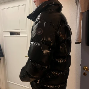 Svart pufferjacka från Napapijri - Säljer en svart, glansig pufferjacka från Napapijri med hög krage och tydlig logga på ärmen. Jackan har dragkedja framtill och två fickor med dragkedja. Perfekt för kalla dagar när du vill hålla stilen och värmen. 