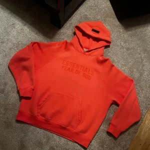 Essentials Fear Of God - *PRIS KAN DISKUTERAS* Ikonisk Essentials Fear of God-hoodien i en stark korall/del av rosa färg. Tillverkad i tjockt, mjukt material med upphöjt ESSENTIALS FEAR OF GOD-tryck framtill. Den har den klassiska oversized-passformen som märket är känt för – avslappnad men lyxig. •	Storlek: [M] •	Färg: Korall •	Skick: Aldrig Använd •	Passform: Oversized, unisex •	Detaljer: Känguruficka, ribbade muddar, logga på bröstet