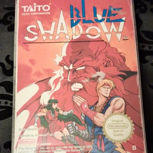 Nintendo Blue Shadow (NES) - Blue Shadow till NES i fint skick, komplett med originalkartong, manual och kassett. Klassisk actionplattformare från Taito med snyggt omslag och tydliga färger. Lättare slitage på framsidan av kartongen, men överlag välbevarat exemplar för samlare.