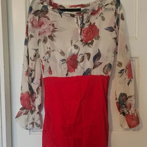 Blommig röd och vit långklänning - Snygg långklänning med blommigt mönster upptill i rött, blått och grönt på vit botten och enfärgad röd nederdel. Långa ärmar med lätt volang vid ärmslut och rund halsringning. Perfekt för dig som vill sticka ut med färg och mönster.