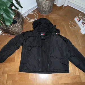 Svart pufferjacka från Tommy Hilfiger - Svart pufferjacka från Tommy Hilfiger med huva och dragkedja. Jackan har klassisk quiltad design, logga på bröstet och ryggen samt långa ärmar. Perfekt för kalla dagar och riktigt snygg streetstil.