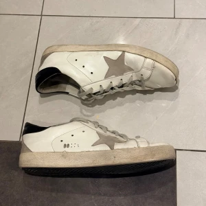Vita sneakers med grå stjärna Golden goose - Säljer ett par vita sneakers från Golden Goose med grå mockadetaljer och en ikonisk stjärna på sidan. Skorna har snörning, svart hälparti och platt sula. Perfekta för dig som gillar en avslappnad och trendig stil.