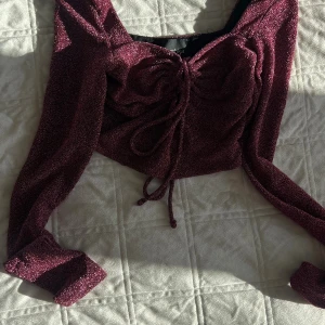 Vinröd glittrig croppad topp Bershka - Säljer en vinröd glittrig croppad topp från Bershka med långa ärmar och snygg snörning framtill. Toppen har en tight passform och är perfekt för att sticka ut med sitt skimrande material och trendiga look.