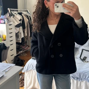 Svart kappa - Säljer en svart skepparkavaj (kappa) med bred krage och dubbelknäppning framtill. Jackan har en klassisk, boxig passform och är tillverkad i ett ulligt material. Perfekt för dig som gillar en stilren och tidlös look.