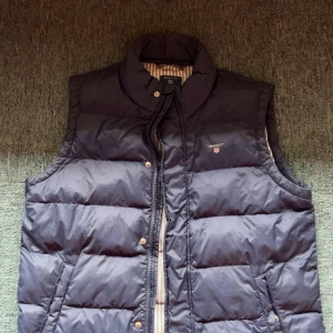 Mörkblå dunväst från GANT 158/164 - Snygg mörkblå dunväst från GANT med quiltad design och ståkrage. Västen har dragkedja och tryckknappar framtill samt två fickor. Insidan är fodrad med beige och blått randigt tyg för extra stilpoäng. Perfekt för lager-på-lager under kyliga dagar. Original pris 2500